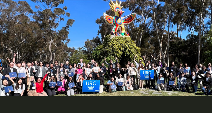 A large group of UC San Diego students pose with their research posters and Undergrad Research Conference Signs, underneath the tall, colorful Sun God statue